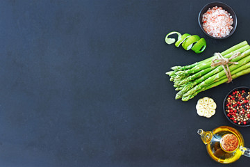Top view of vegetarian cooking background. Bunch of green asparagus, lime skin, ground pepper, garlic, olive oil and himalayan salt on a dark background with a space for text.