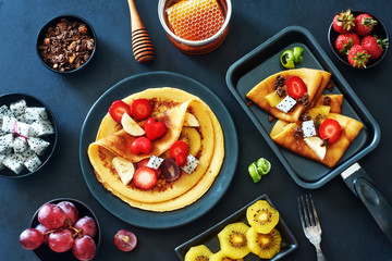 Homemade crepes with strawberries, honey, chocolate granola and exotic fruit. Delicious breakfast with thin pancakes on a black background. 