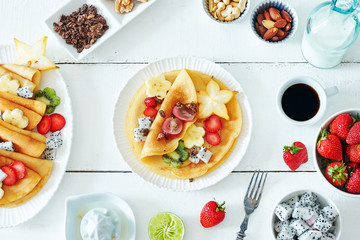 Top view of thin pancakes with starfruit, banana, kiwi, grape, strawberry and honey. Milk and coffee on a white table. Healthy breakfast with crepes. 
