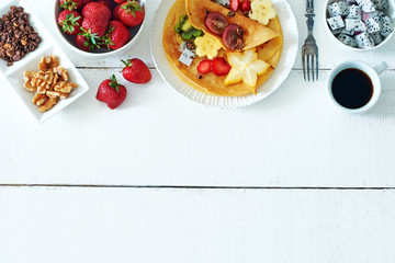 Top view of homemade crepes with exotic fruits, granola and black coffee over white background with a copy space. 