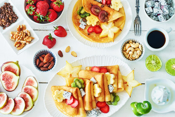 Healthy breakfast with thin pancakes, exotic fruits (starfruit, pitaya, banana, kiwi), strawberry, figs, granola, coffee and milk on a white table. 