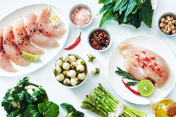 Raw chicken meat on a white plates, fresh greens and vegetables, salt, pepper and olive oil on a white table. Light dinner ingredients. 