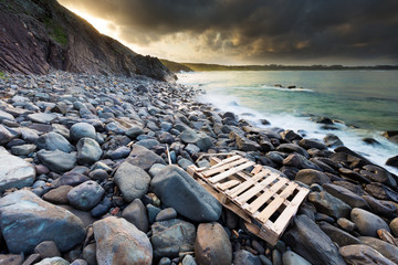 A broken driftwood packing crate washed ashore on a boulder strewn coastline under  illuminated...