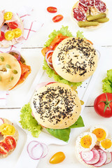 Vertical top view of assorted bagels with salami, ham, egg, smoked salmon, cream cheese and pesto, vegetables on white table. Healthy picnic concept. 
