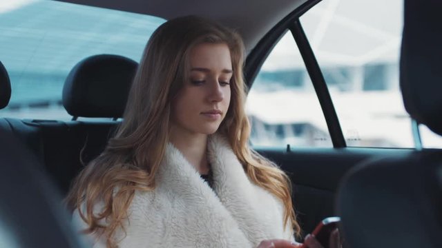 Young pretty blonde driving in the car typing on her smartphone communicating with friend of her.