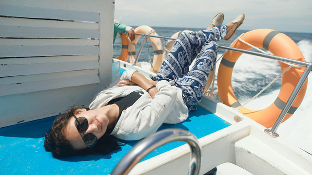 Young cheerful girl is having cruise on board of a yacht in the open ocean. Smiling woman is lying on deck of a little ship in the sea with her legs in the air between two orange life-buoy rings.