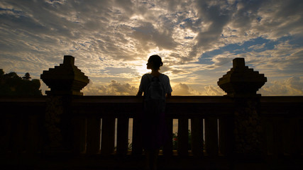 The girl is not tall with a backpack standing on the porch near the column. She travels in Bali. She enjoys beautiful views and sunsets. Bali amazing and fascinating place.