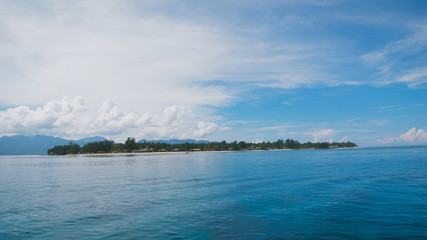 View from a boat on a small island with lots of trees and greenery, located in a blue ocean, over there is a clear blue sky with clouds