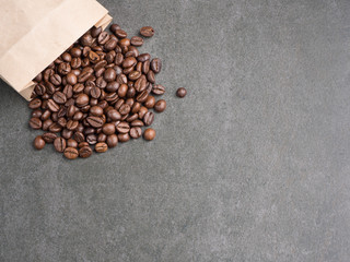Roasted coffee beans scattered from paper bag over dark gray granite plate, captured from top view with sharp focus