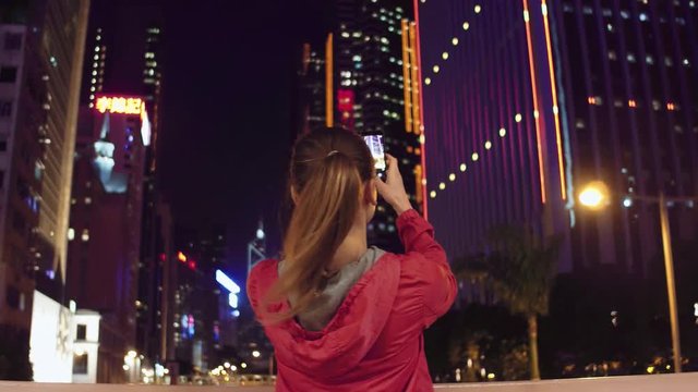 Young woman taking photos of buildings on her phone at night, Hong Kong.