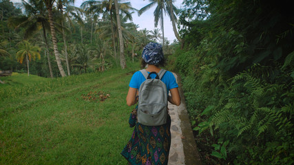 Young woman with a backpack walking along the stone footpath in the green of the jungle. Tourist is actively pursuing their vacation on a tropical island.