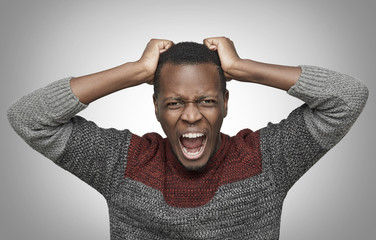 Mad African American man shouting with mouth wide open and closed eyes, holding hands on his head,...