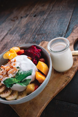 The plate of exotic fruits ( mango, banana, dragon fruit and papaya) and yogurt on top in the bowl on the wooden table with cocnut milk and spoon, Indonesia, Bali
