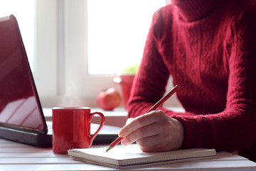 cozy working atmosphere in red tones/ person writes in a notebook next to a red mug and a laptop, sitting in front of the window