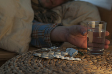 Ein Mann liegt mit Erkaeltung im Bett / Ein Mann liegt mit Erkaeltung im Bett und greift nach ein Glass Wasser, vor ihm liegen Medikamente, geringe Tiefenschaerfe.
