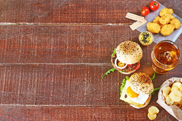 Glass of wheat beer and snacks: homemade beef burgers, chicken nuggets with cheese sauce, crispy potato chips, cherry tomatoes over dark wooden background. Copy space.