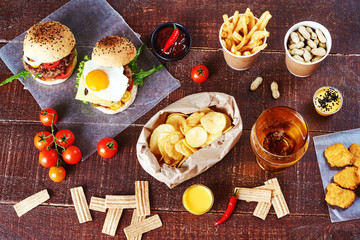 Wheat beer and set of snacks on a dark wooden table: beef burgers, potato chips, chicken nuggets, peanuts, fresh tomatoes. Junk food set. Top view.