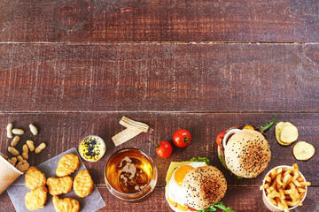 Glass of beer and snack variety over dark wooden background. Beef burgers, chicken nuggets, peanuts, french fries with cheese sauce, crispy potato chips, bbq sauce, fresh tomatoes. Copy space.