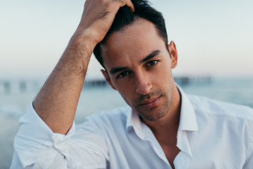 Close up portrait of handsome man wearing white shirt at the sea background. Travel vacation holiday. Attractive man looking front with hand on hair. Fashion caucasian man model elegant outside.