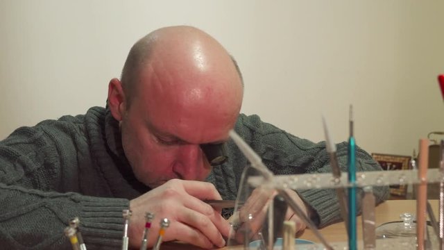 Mature male clockmaker beginning his work. Caucasian man putting on the loupe on his eye. Bald clock repairer taking screwdriver from his work bench