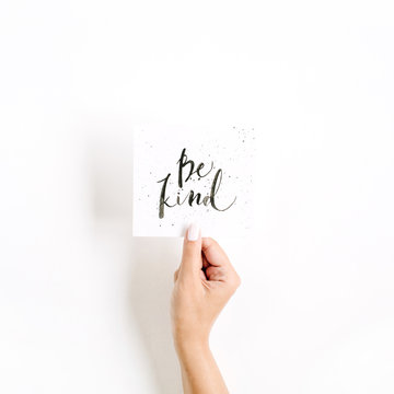 Minimal pale composition with girl's hand holding card with quote Be Kind written in calligraphic style on paper on white background. Flat lay, top view