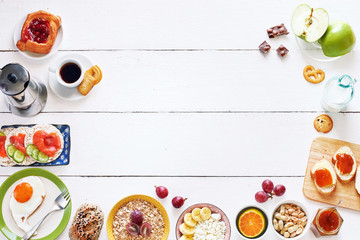 Breakfast menu food frame with a copy space. Eggs, croissants, oatmeal with banana, cereal, danish pastry, crisps with salmon, apricot jam, fresh bread, chocolate, coffee and milk on a white table.