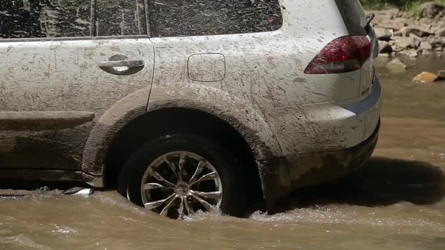 Dirty and mud-splattered white car crosses a river and travels on river stones. Camera shoots from a close wheel angle.