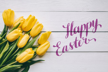 Happy easter hand lettering calligraphy text. Yellow tulips bunch on white wooden planks rustic barn rural table background. Letters, inscription. Beautiful horizontal flat lay postcard.