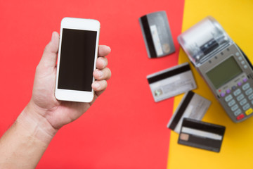 Human hand holding Smartphone for payment with blurred credit card and credit card machine on red and yellow background.