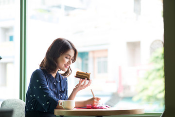 Young beautiful woman in the cafe near the window, thinking and writing something with cake in hand.