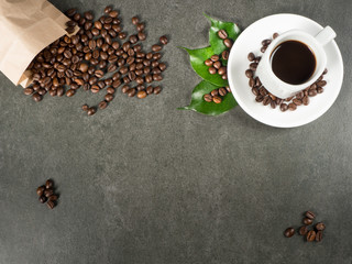 White cup filled with hot coffee, roasted beans and green leaves scattered from paper bag. Dark gray granite for background, captured from top view with sharp focus