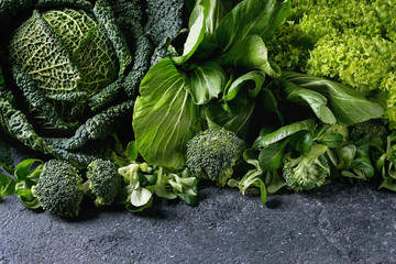 Variety of raw green vegetables salads, lettuce, bok choy, corn, broccoli, savoy cabbage as frame over black stone texture background. Space for text - obrazy, fototapety, plakaty