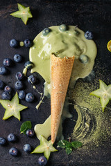 Waffle cone of melting Green tea matcha soft elastic ice cream with mint leaves, carambola, lime, blueberries over black texture background. Top view.