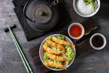 Fried tempura shrimps on lettuce salad with sauces. Served in traditional china plate with chopsticks on wood serving board. Teapot, bowl of rice. Over old metal background. Top view. Asian dinner
