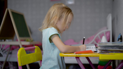 Little girl is drawing the picture with pencils in studio sitting at the table. Small child is learning to paint with soft-tip pens of different colours. Cute kid crayoning the pattern with pleasure.