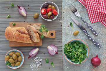 Green and black olives, loaf of fresh multigrain bread, corn salad and red onion over old wood background.