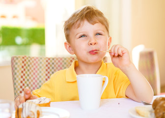 Happy kid eats his dessert while sitting at a table on a summer terrace lit with bright daylight. Boy looks away. He dressed in casual yellow polo.