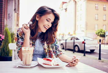 Happy young woman eating cake at outdoors cafe and read message on her smart phone
