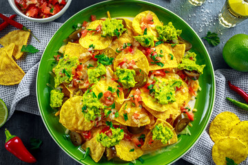 Nachos with cheese, jalapeno peppers, red onion, parsley, tomato, salsa, guacamole sauce and tequila on green plate.