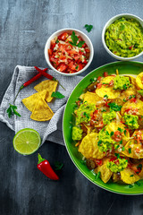 Nachos with cheese, jalapeno peppers, red onion, parsley, tomato, salsa, guacamole sauce and tequila on green plate.