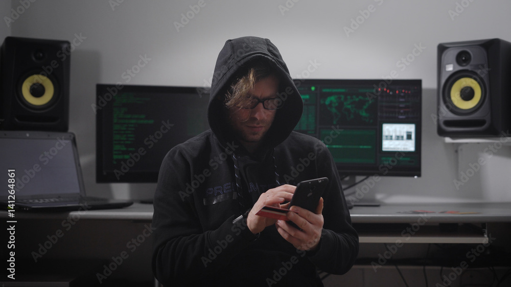 Poster close up of a hooded hacker making cyber attack on bank network with the help of smartphone in his h