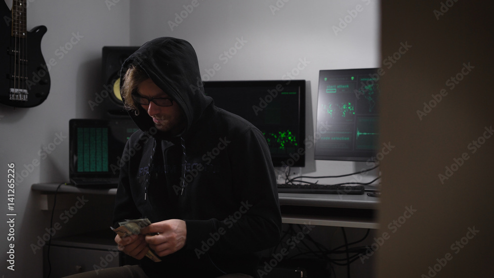 Poster a hacker in the computer room. a man in a jacket with a hood, glasses and money in hands sitting in 