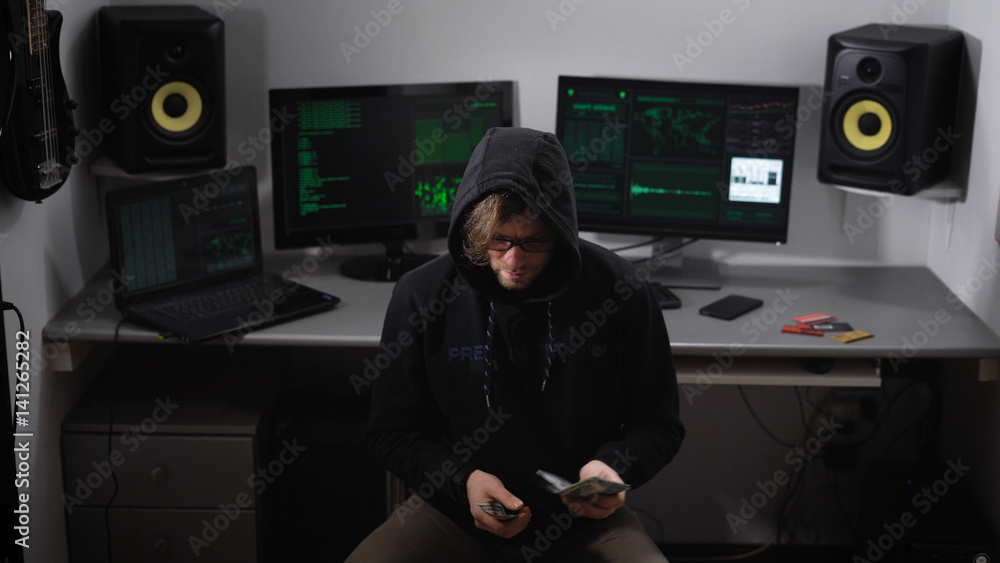 Poster hooded hacker in black clothes sitting on a chair in the room and counting prepayment for hacking ne