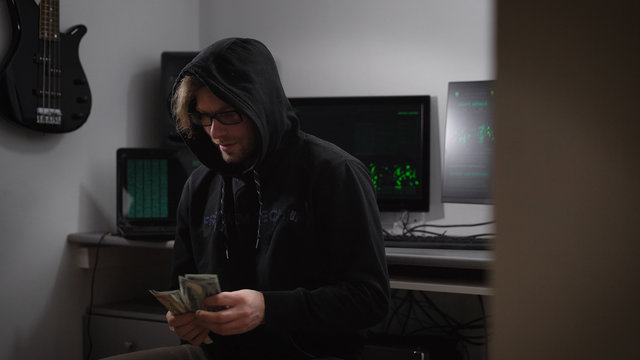 Professional hacker consider his money is sometimes dissatisfied looking at the camera and then lays down a stack in pocket of sweater. In the background are two monitor that handles electronic data.