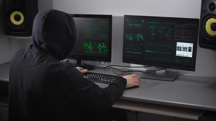 Back view of a hooded angry hacker in black making cyber attack on bank network and failuring the process. Aggravated man is throwing keyboard on the table and striking it with his fists.