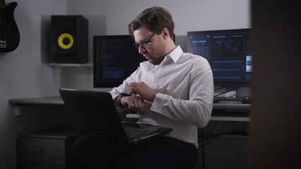 Young programmer in a white shirt and stylish glasses working on a laptop that is on his knees on the background several monitors with more information.