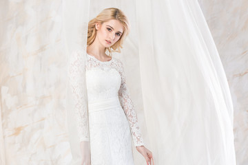 Fototapeta premium Portrait of the gorgeous thoughtful blonde bride with beautiful curly hair in a white wedding lacy dress. beautiful young model with bridal hairstyle and makeup in a wedding dress.