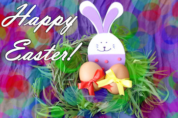 easter hand made decorated greeting card: yellow eggs and hand made festive plastic foam bunny in green grass twigs nest on purple background with inscription happy easter