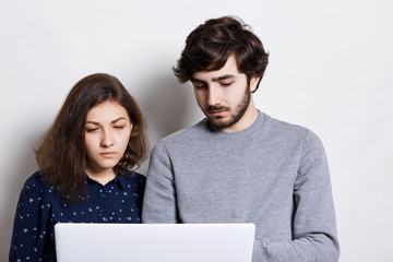 Stylish bearded man and beautiful woman standing close to each other enjoying modern technologies, using high-speed internet connection and laptop, browsing social media .
