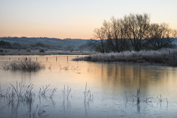 Beautiful vibrant English countryside lake image with frost and frozen lake in Winter at sunrise